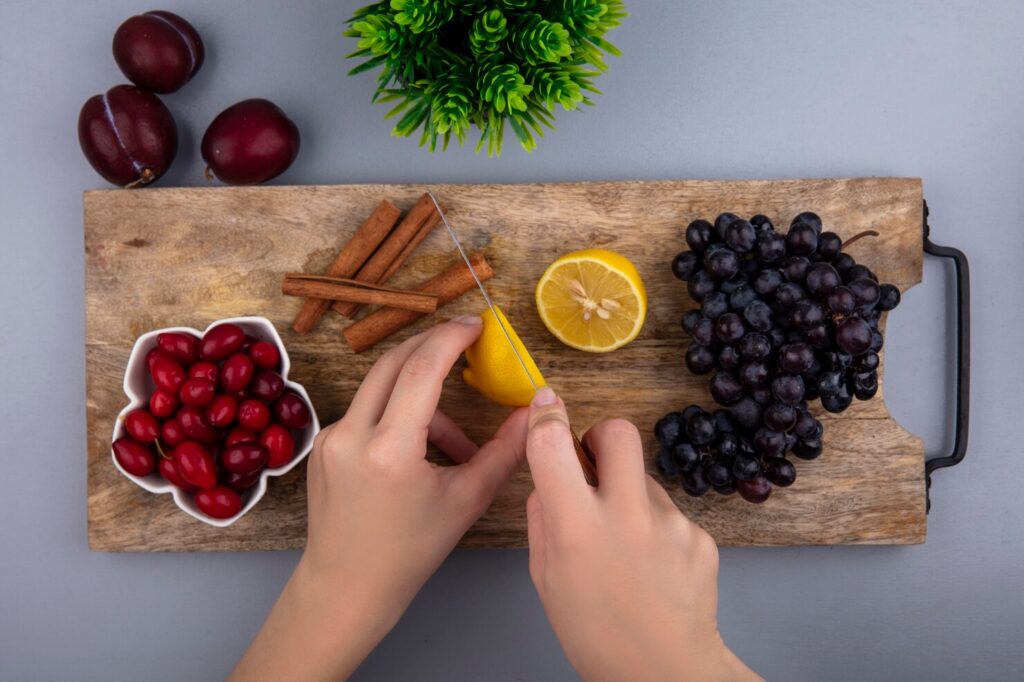 top-view-female-hands-cutting-lemon-with-knife-cornel-berries-grape-cinnamon-cutting-board-pluots-plant-gray