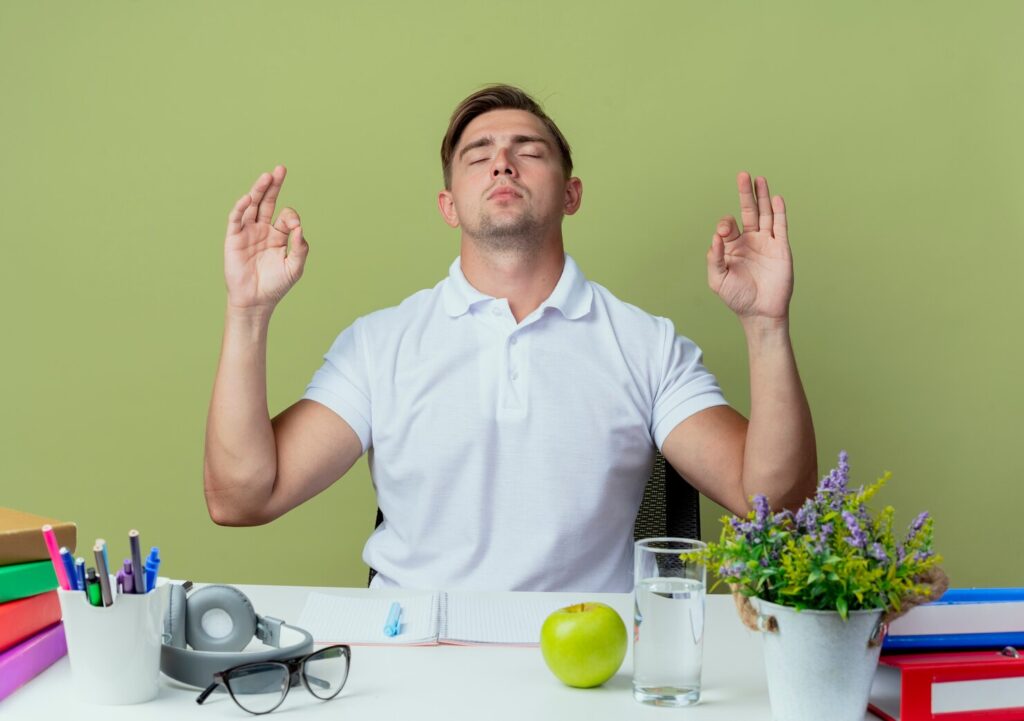 with-closed-eyes-young-handsome-male-student-sitting-desk-with-school-tools-showing-meditation-gesture-isolated-olive-green